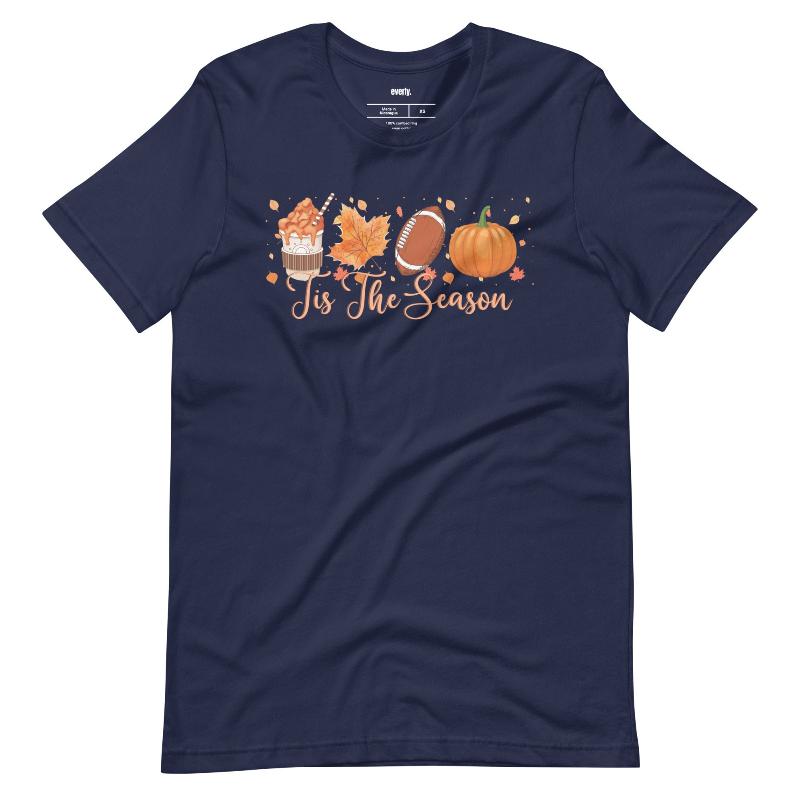  'Tis The Season' design with fall-themed graphics including a cupcake, maple leaf, football, and pumpkin, surrounded by falling leaves on a navy shirt. 