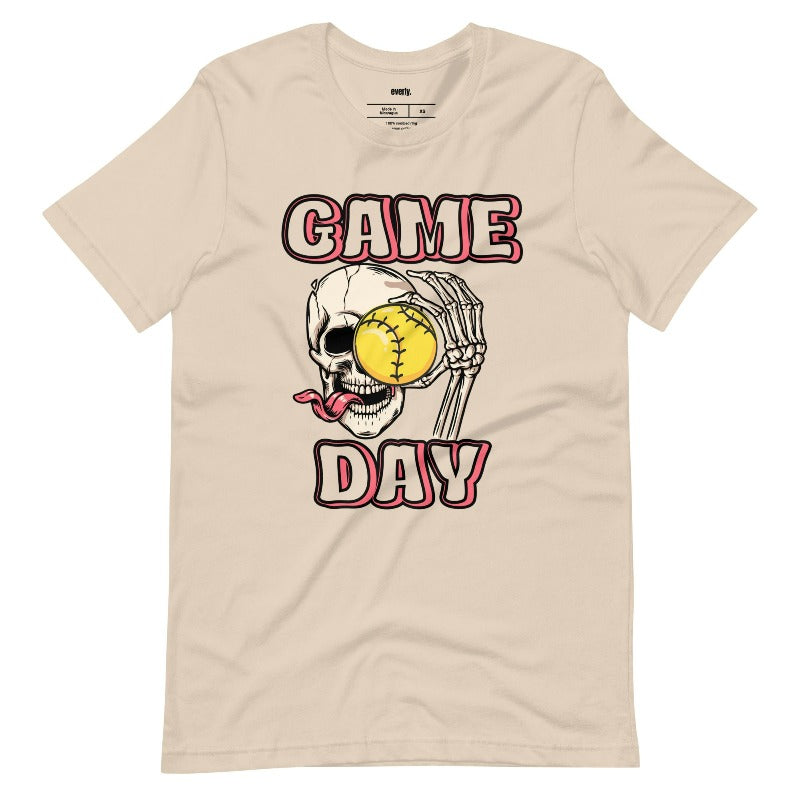 Softball game day skeleton skull holding a softball on a soft cream graphic tee.