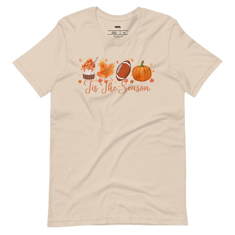  'Tis The Season' design with fall-themed graphics including a cupcake, maple leaf, football, and pumpkin, surrounded by falling leaves on a beige shirt. 