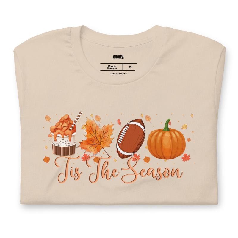  'Tis The Season' design with fall-themed graphics including a cupcake, maple leaf, football, and pumpkin, surrounded by falling leaves on a beige shirt. 