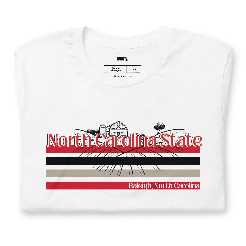 A vibrant NC State design with "North Carolina State" written in red across the top, stripes in red, black, and white, and "Raleigh, North Carolina" at the bottom on a white graphic tee.