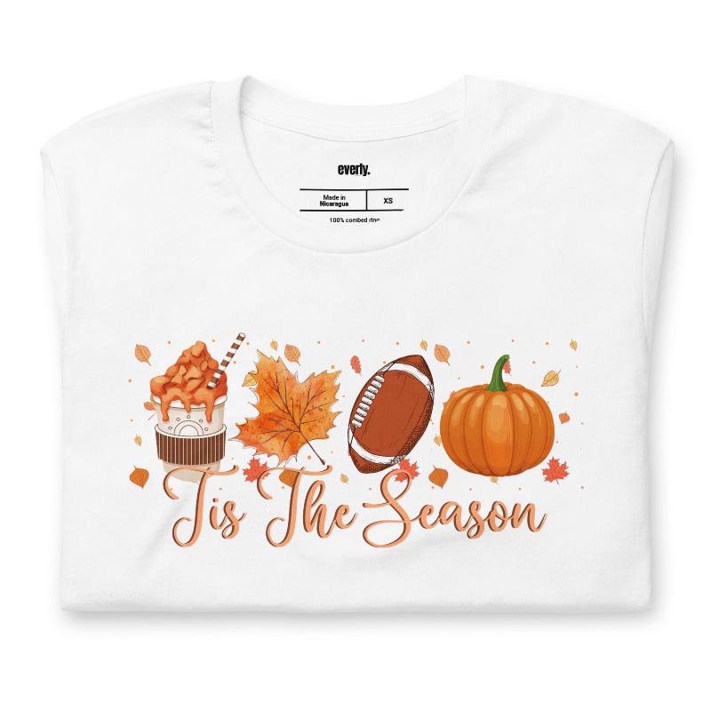 'Tis The Season' design with fall-themed graphics including a cupcake, maple leaf, football, and pumpkin, surrounded by falling leaves on a white shirt. 